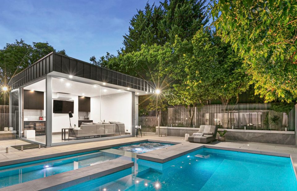 Poolside pavilion with outdoor kitchen lit at dusk beside the pool and spa