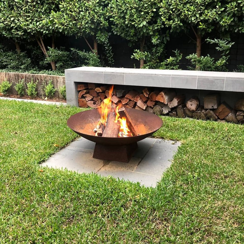 Corten fire bowl burning on a bluestone pad in the lawn, firewood stored beneath a honed bluestone bench